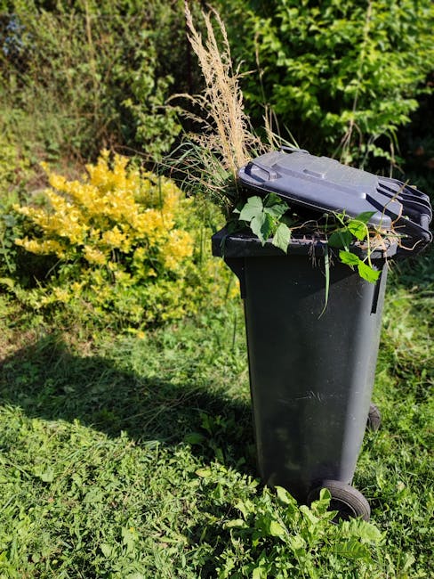 A black wheeled rubbish bin positioned on a grassy outdoor area, filled with garden waste including dried grasses, leafy branches, and other organic plant debris. The bin is situated near a lush green
